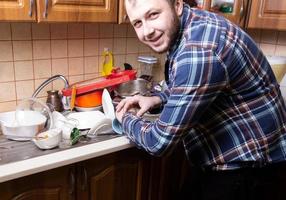 een jonge, bebaarde man kijkt op zijn horloge en is blij met de hoeveelheid vuile vaat die in de gootsteen ligt foto