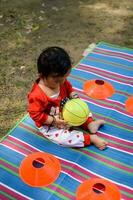 schattig weinig Indisch zuigeling zittend genieten van buitenshuis schieten Bij maatschappij park in Delhi, schattig baby jongen zittend Aan kleurrijk mat met gras in de omgeving van, baby jongen buitenshuis schieten foto