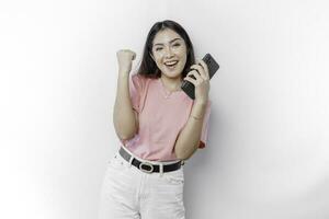 een jong Aziatisch vrouw met een gelukkig geslaagd uitdrukking vervelend roze t-shirt en Holding smartphone geïsoleerd door wit achtergrond foto