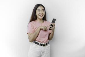 een portret van een gelukkig Aziatisch vrouw is vervelend roze t-shirt en Holding haar telefoon, geïsoleerd door wit achtergrond foto