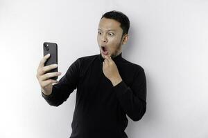 geschokt Aziatisch Mens vervelend zwart overhemd Holding zijn telefoon met zijn mond breed open, geïsoleerd door wit achtergrond foto