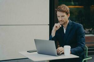 knappe peinzende jongeman die op laptop op internet surft terwijl hij op afstand in een café op de stoep werkt foto