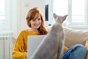 jong mooi vrouw aan het doen online werk Bij huis, mooi sphynx kat afleidend eigenaar van werk foto