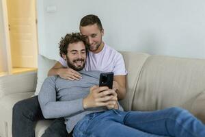 schattig jong homo paar video roeping hun vrienden in hun leven kamer Bij huis. twee mannetje geliefden glimlachen vrolijk terwijl groet hun vrienden Aan een smartphone. jong homo paar zittend samen. foto