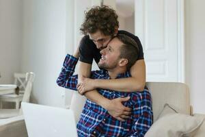 lief homo paar lachend samen terwijl zittend in hun leven kamer Bij huis. twee romantisch jong mannetje geliefden hebben pret surfing de internet binnenshuis. jong homo paar leven samen. foto