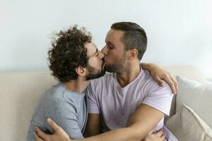 lief homo paar zoenen terwijl zittend in hun leven kamer Bij huis. twee romantisch jong mannetje geliefden hebben pret binnenshuis. jong homo paar leven samen. foto