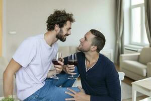 lgbtq paar omarmen elk andere en slinkend wijn binnenshuis. twee romantisch jong mannetje geliefden op zoek Bij elk andere terwijl zittend samen in hun leven kamer. jong homo paar wezen romantisch Bij huis. foto