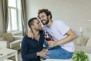 lgbtq paar omarmen elk andere en slinkend wijn binnenshuis. twee romantisch jong mannetje geliefden op zoek Bij elk andere terwijl zittend samen in hun leven kamer. jong homo paar wezen romantisch Bij huis. foto