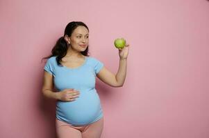 mooi aantrekkingskracht verwachtend vrouw, zetten hand- Aan haar zwanger buik, Holding een groen appel Aan geïsoleerd roze achtergrond foto