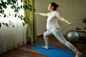 zwanger vrouw beoefenen yoga, staand Aan geschiktheid mat in krijger houding, genieten van gezond levensstijl gedurende haar zwangerschap foto