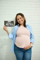 gelukkig positief volwassen zwanger vrouw, strelen haar buik, lachend, tonen Bij camera de echografie van haar baby in baarmoeder foto