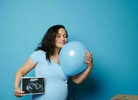 verrukkelijk zwanger vrouw knuffelen blauw ballon, poseren met echografie beeld van haar toekomst baby jongen, blauw backdrop foto