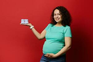 verwachtend zwanger moeder, gravid vrouw Holding schattig blauw gebreid baby slofjes, geïsoleerd rood achtergrond. kopiëren ruimte. foto