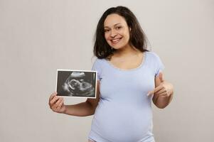 gelukkig zwanger verwachtend moeder Holding scannen van haar kind in baarmoeder, glimlachen Bij camera en richten vinger Bij haar buik foto