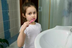 tandheelkundig zorg en mondeling hygiëne voor gezond wit baby tanden. aanbiddelijk weinig kind meisje poetsen tanden, op zoek Bij camera. foto
