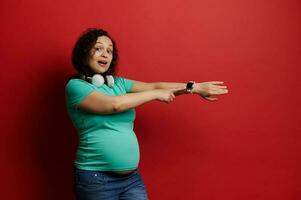 mooi zwanger vrouw met koptelefoon, tonen Bij camera haar slim pols horloge, rood achtergrond. zwangerschap mobiel app foto