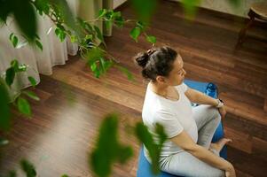 zwanger vrouw werken uit in leven kamer interieur, zittend in prenataal gemakkelijk lotus houding, bereiken harmonie, lichaam balans foto