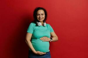 mooi zwanger vrouw met handen Aan haar buik, gevoel gelukkig door bewegingen van haar baby in baarmoeder, geïsoleerd rood backdrop foto