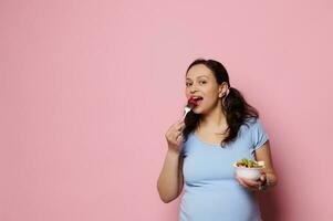geïsoleerd portret Aan roze achtergrond van aantrekkingskracht charmant zwanger verwachtend vrouw aan het eten fruit salade, glimlachen Bij camera. foto