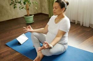 mooi zwanger vrouw aan het doen zwangerschap yoga en adem opdrachten Bij huis, zittend in lotus positie mediteren Bij huis foto