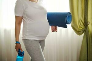 buik van zwanger vrouw in laat zwangerschap, in wit mockup t-shirt, Holding oefening mat, klaar voor geschiktheid en yoga foto