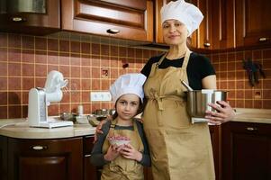 gelukkig zwanger vrouw en haar lief dochter in schort en chef-kok hoed, genieten van Koken samen in de knus huis keuken foto