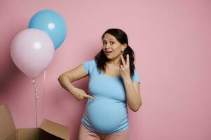 multi etnisch aantrekkingskracht zwanger vrouw in blauw t-shirt, ervan uitgaand Tweelingen, geïsoleerd roze achtergrond met roze blauw ballonnen foto