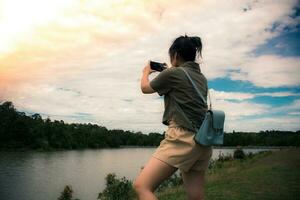 jong Aziatisch meisje naar reizen de vakantie. ze was vervelend een groen overhemd - schouder tas, op zoek Bij de zonsondergang en gebruik makend van mobiel telefoon naar nemen afbeeldingen van rivier- en bergen of video telefoontje voelen ontspannen. foto