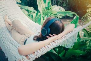 jong ontspannen vrouw luisteren naar muziek- met hoofdtelefoons Aan wit hangmat in tuin Bij huis. ochtend- zon. langzaam leven, weekend vrije tijd werkzaamheid. quarantaine en zelf isolatie periode. foto