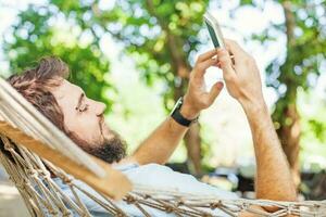 detailopname van een Kaukasisch Mens gebruik makend van mobiel telefoon wit swinging in een hangmat foto