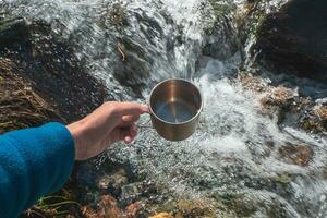 mok met zuiver glaciaal water van een berg stroom. hand- Holding een wandelaars kop met schoon drinken water. foto