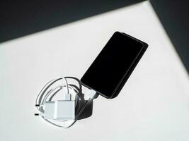 een zwart smartphone met een opladen draad Aan een wit tafel met schaduwen. foto
