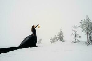 jong mooi vrouw in lang zwart jurk met oud lantaarn over- winter heuvel achtergrond en sneeuwval. fee verhaal meisje Aan polair winter landschap. zwart heks in de sneeuw. foto