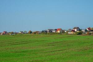 een groen voorjaar veld- in voorkant van een modern dorp Aan een heuvel tegen een Doorzichtig blauw lucht. agrarisch land. foto