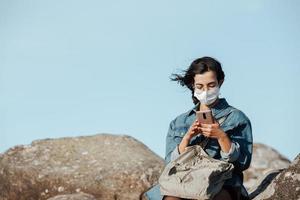een jonge vrouw die een masker draagt en zijn telefoon controleert terwijl de wind met exemplaarruimte waait foto
