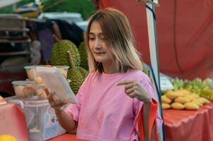 Aziatisch vrouw staand Bij de lokaal markt en Holding een fruit voor kiezen buying fruit naar bereiden voor diner, biologisch gezond voedsel lokaal markt concept foto