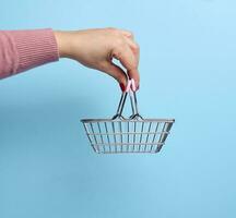 vrouw hand- Holding een miniatuur metaal boodschappen doen mand Aan een blauw achtergrond foto