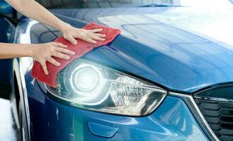 auto wassen onderhoud. Mens hand- Holding rood microfiber kleding en Pools koplamp van blauw auto. auto zorg onderhoud bedrijf concept. Mens schoonmaak en detaillering luxe auto. schoon water druppels van luxe auto. foto