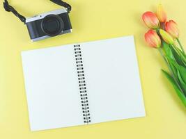 vlak leggen van blanco bladzijde geopend notitieboekje, digitaal camera en rood geel tulpen bloemen Aan geel achtergrond met kopiëren ruimte. foto