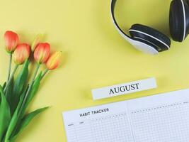 vlak leggen van gewoonte tracker boek met houten kalender augustus, hoofdtelefoons en tulpen Aan geel achtergrond. foto