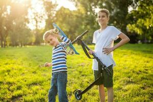 twee jongens zijn hebben pret met skateboard en scooter in park. speels kinderen in park, gelukkig jeugd. foto