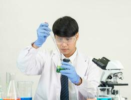 Aziatisch mannetje leerling wetenschapper vervelend een dokter japon in de laboratorium, op zoek Bij de resultaten door een microscoop. Aan een tafel in een wetenschappelijk Onderzoek laboratorium met test buizen Aan een wit tafel foto