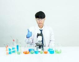 Aziatisch mannetje leerling wetenschapper vervelend een dokter japon in de laboratorium, op zoek Bij de resultaten door een microscoop. Aan een tafel in een wetenschappelijk Onderzoek laboratorium met test buizen Aan een wit tafel foto