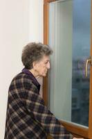 oud vrouw van 80 jaren oud blijft dichtbij naar de venster foto