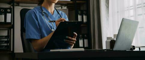 echt vrouw dokter gebruik makend van laptop en schrijven aantekeningen in medisch logboek zittend Bij bureau. jong vrouw professioneel medisch arts vervelend wit jas en stethoscoop werken Aan computer Bij werkplek. foto