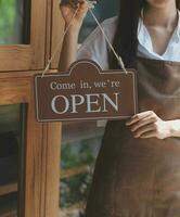 Welkom. open. barista, serveerster vrouw draaien Open teken bord Aan glas deur in modern cafe koffie winkel klaar naar onderhoud, cafe restaurant, kleinhandel op te slaan, klein bedrijf baasje, voedsel en drinken concept foto