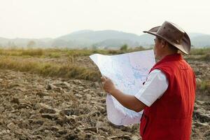 Aziatisch Mens ontdekkingsreiziger draagt hoed, rood hesje shirt, houdt kaart naar onderzoeken land- grens. concept, land- planning, verkennen eigendom. geodetisch enquête Oppervlakte. schat jacht- Aan land. avontuur levensstijl. foto