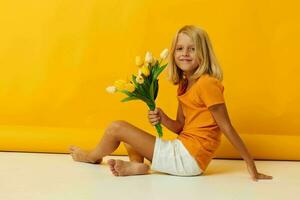 weinig meisje zittend Aan de verdieping met een boeket van geel bloemen pret geel achtergrond foto