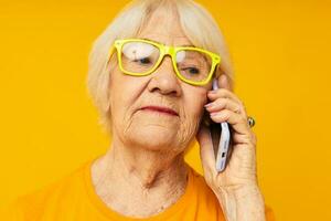 portret van een oud vriendelijk vrouw pratend Aan de telefoon in geel bril geïsoleerd achtergrond foto