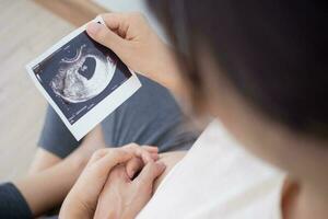 zwanger vrouw is op zoek Bij een echografie foto van foetus. moeder voorzichtig accenten de baby Aan maag. Vrolijk, familie, groei, zwangerschap, genot , bereiden pasgeboren, nemen zorg, gezondheidszorg, buik.